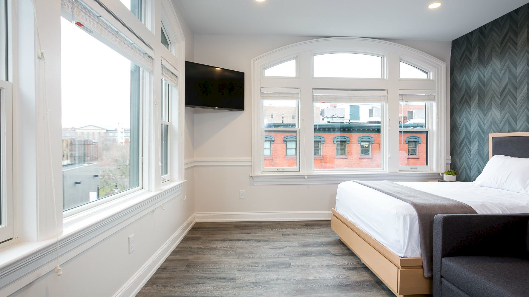 A modern bedroom with large windows, a mounted TV, a bed with white bedding, grey walls, and a stylish dark accent wall with a chevron pattern.