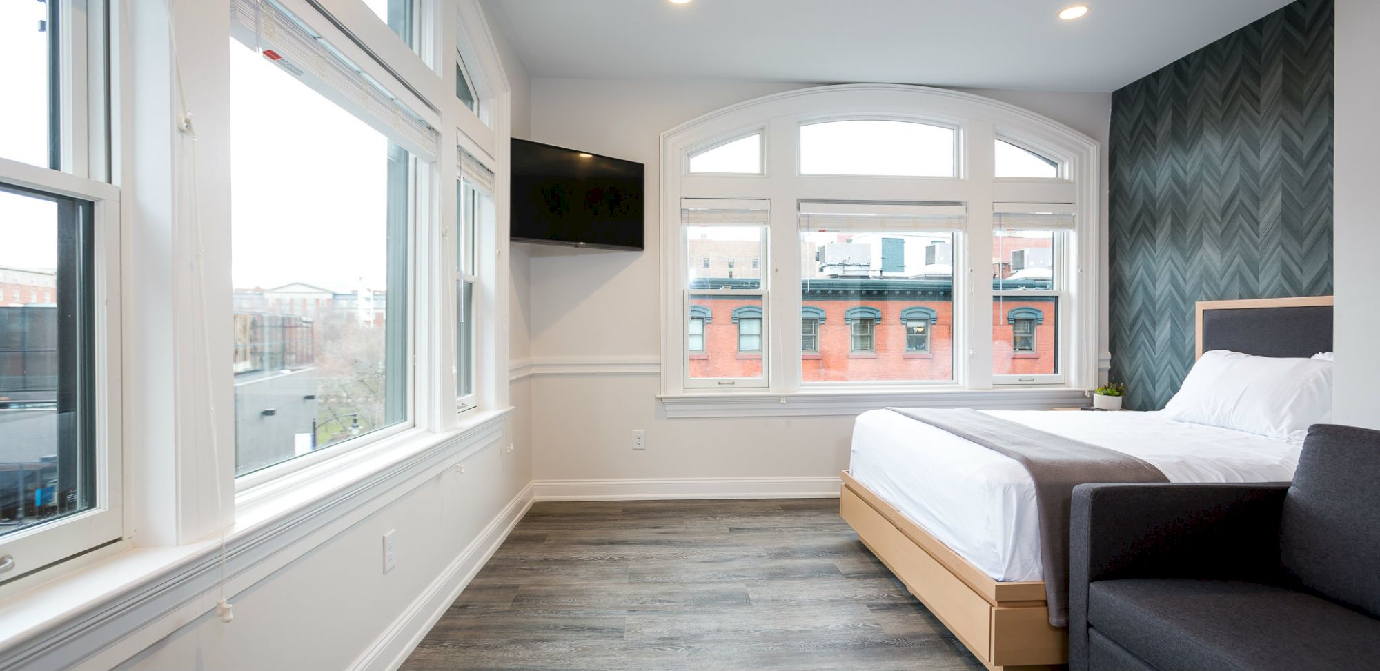 A modern bedroom with large windows, a mounted TV, a bed with white bedding, grey walls, and a stylish dark accent wall with a chevron pattern.