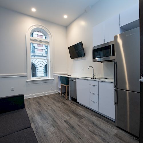 A compact modern kitchen with white cabinets, stainless steel appliances, a wall-mounted TV, and a black sofa by two arched windows.