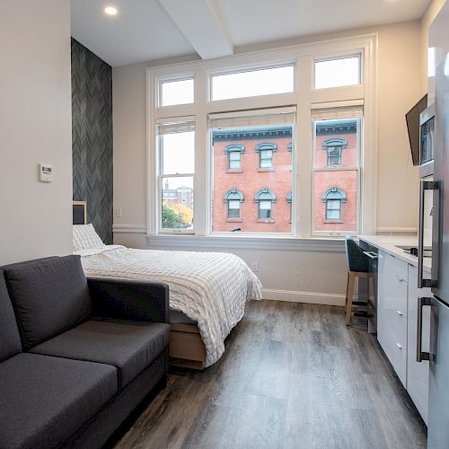 A modern studio apartment features a cozy bed, a couch, a kitchen area, and large windows overlooking a building with red brick exterior.