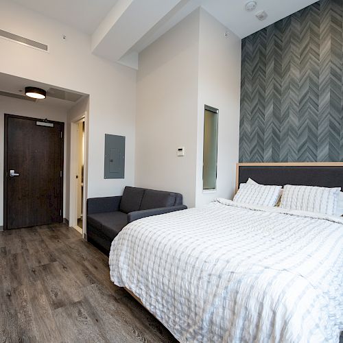 The image shows a modern bedroom with a large bed, a nightstand, a small couch, and a dark wooden door, featuring a chevron-patterned accent wall.