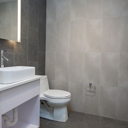 A modern bathroom with a sink, mirror, and toilet, featuring gray tiled walls and floor.