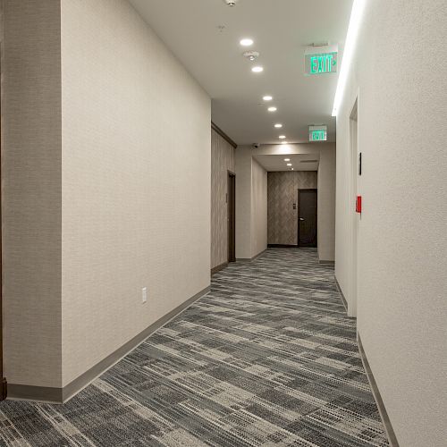 A well-lit hallway with patterned carpet, beige walls, ceiling lights, and exit signs. There's a fire alarm on the right wall.