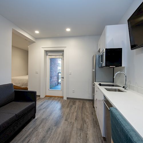 A modern studio apartment features a sofa, kitchenette, wall-mounted TV, and a bed area with wooden floors and bright lighting.