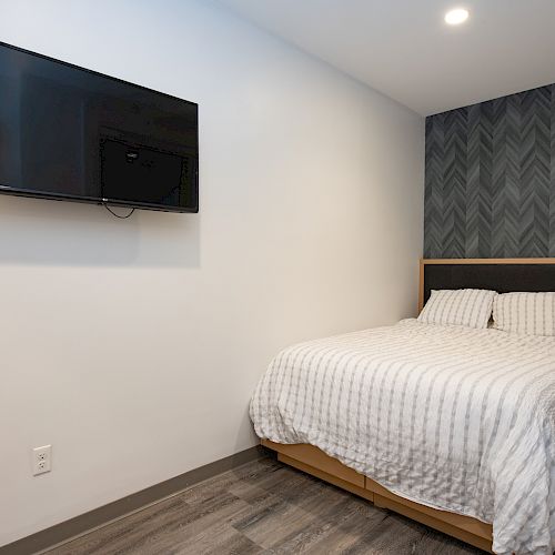 A modern bedroom with a wall-mounted TV, a bed with striped bedding, and wooden furniture, against a dark patterned accent wall.