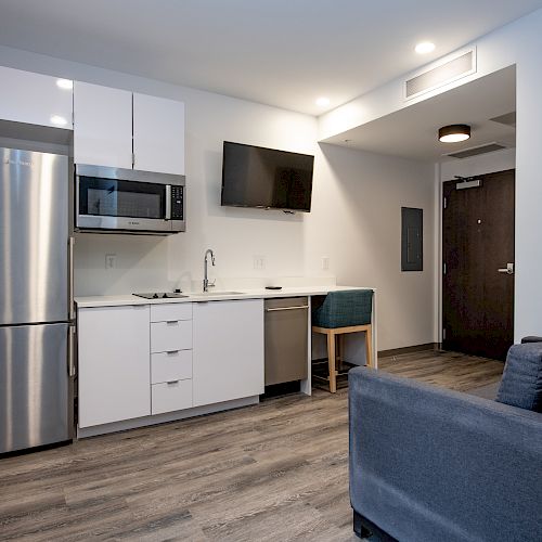 A small modern kitchen and living area with a refrigerator, microwave, sink, TV, chair, and couch, featuring light flooring and white cabinets.