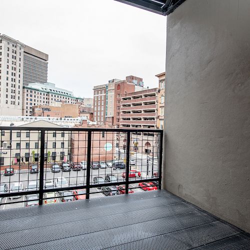 A balcony with a metal floor and railing overlooks a city street filled with parked cars and various buildings.