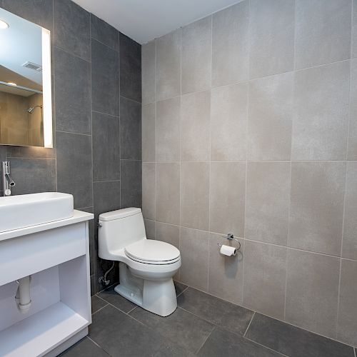 A modern bathroom with a white sink and vanity, toilet, toilet paper holder, and large gray tiles on the walls and floor.