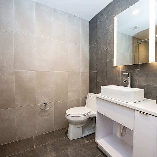 A modern bathroom with gray tiles, a toilet, a white vanity with a vessel sink, and a mirror with lighting.