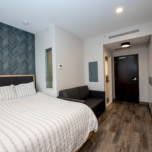 A modern studio apartment features a bed, gray couch, kitchenette, and a wooden floor. The background includes patterned wallpaper and a front door.