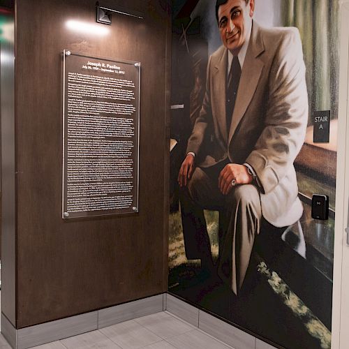 The image shows a wall mural of a man in a suit next to a plaque with text, illuminated by a light. The setting appears to be indoors.