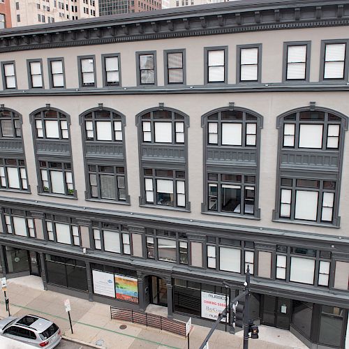 The image shows a multi-story building with a grey facade and black window frames situated on a corner in an urban area.