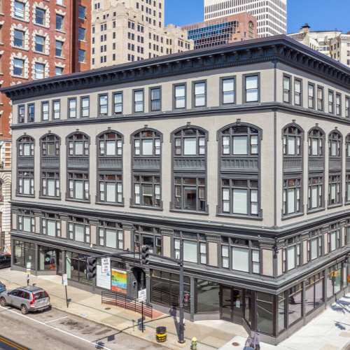 A multi-story historic building on a city street corner, featuring arched windows and ground-level storefronts, with surrounding cars and nearby buildings.