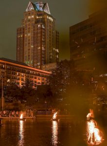 The image depicts a waterfront scene at night with lit fire installations on the water, and a tall illuminated building in the background.