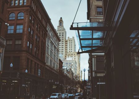 The image depicts an urban street scene with tall buildings on both sides and a prominent skyscraper in the background under a cloudy sky.