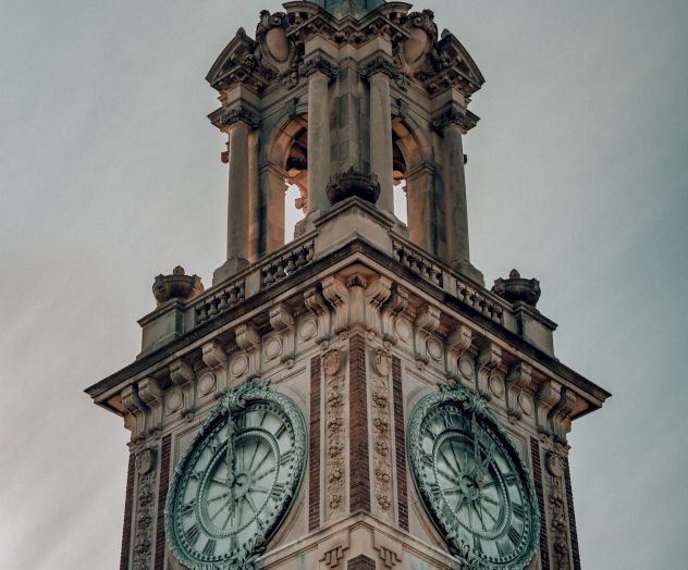 A tall, ornate clock tower with large clock faces on two sides, set against a cloudy sky. The architectural design appears historic and detailed.