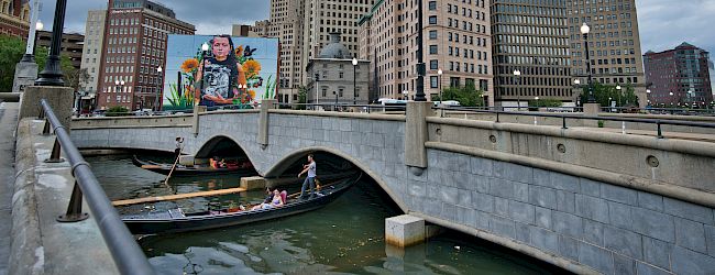 The image shows a bridge over a waterway with people in boats, surrounded by tall buildings, and a mural on one of the structures in an urban setting.