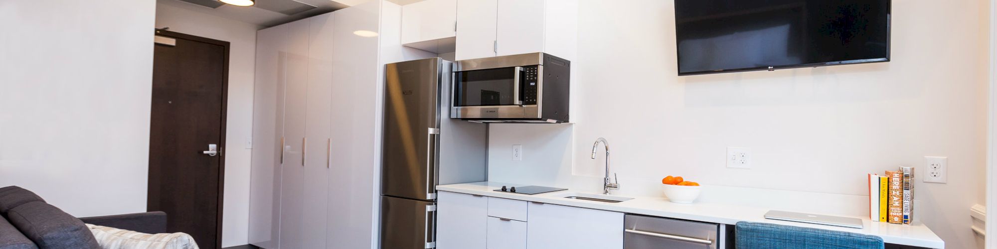 A modern studio apartment with a compact kitchen featuring stainless steel appliances, a wall-mounted TV, a sofa, and a cozy bed.