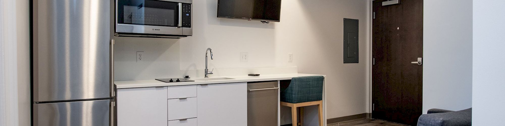 A compact modern kitchenette with stainless fridge, microwave, white cabinets, small sink, toaster, and a wall-mounted TV, plus a blue sofa in a cozy studio.